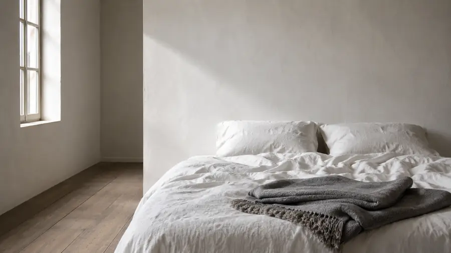 Bed met linnen beddengoed en een gevouwen wollen deken in een lichte kamer met kalkwanden.